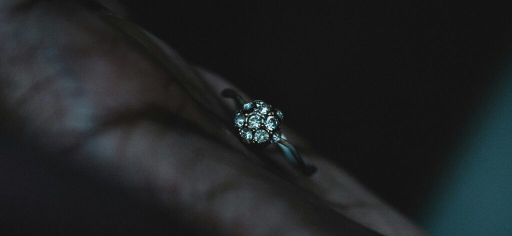 A close-up photograph of a delicate diamond ring resting on a hand. The ring features a floral cluster setting made of multiple small round diamonds arranged in a circular pattern, shining softly in the dim, moody lighting.